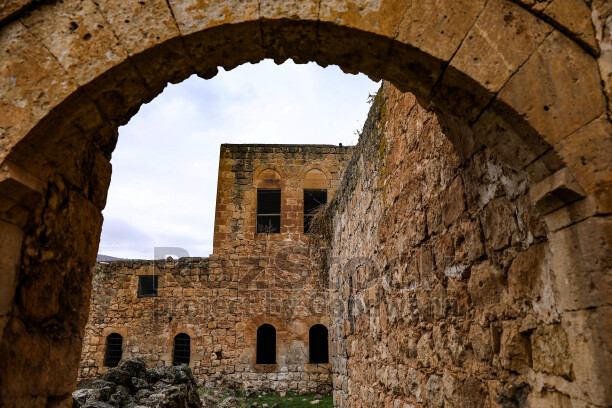 Mardin Savur'da tarihi taş yapıların arasında yer alan kemerli bir geçitten görülen eski yapı dokusu. Sarı kalker taşından örülen duvarlar, kemerli pencereler ve yıpranmış yüzeyler bölgenin geleneksel mimarisini yansıtıyor.